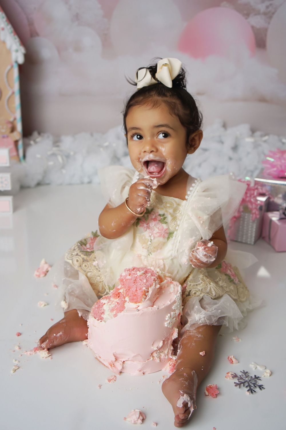 Joyful toddler girl in a cream tulle dress with a bow enjoys her pink smash cake at a winter-themed first birthday session.