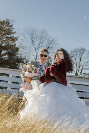 A newlywed couple sits together in tall grass wearing a white dress and burgundy wrap.