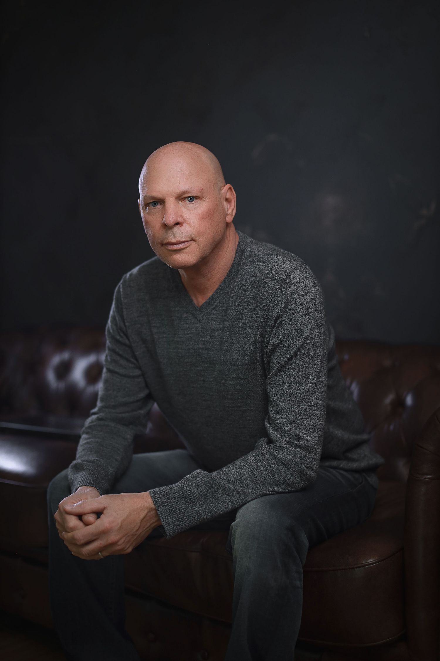 Professional studio portrait of a person in grey sweater seated on leather couch against dark background.