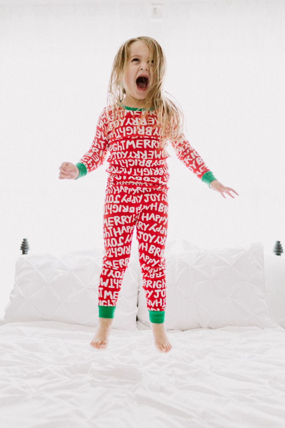 Child wearing red and white holiday pajamas jumping excitedly on a white bed.