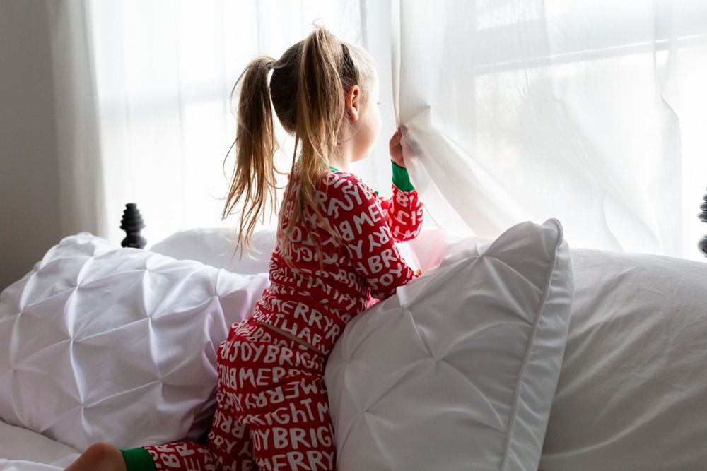 Child in red holiday pajamas looking out window while kneeling on white bedding.
