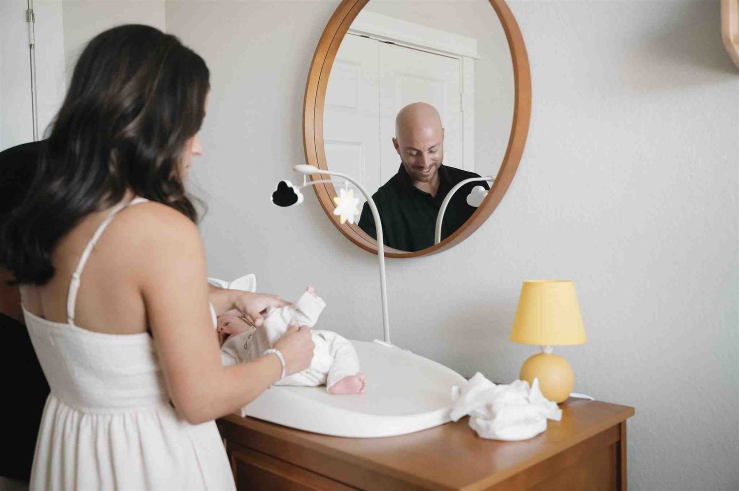 A figure in a white dress folds clothes at a dresser while another person is reflected in a round mirror on the wall.