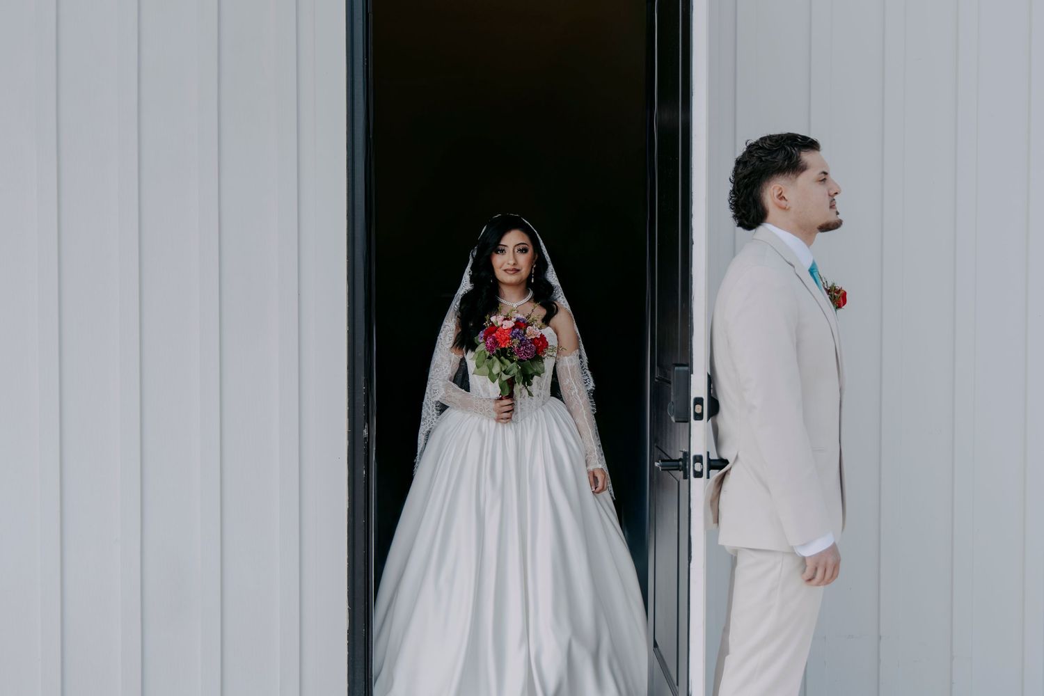 Bride in white ball gown holding bouquet standing in doorway with groom outside.