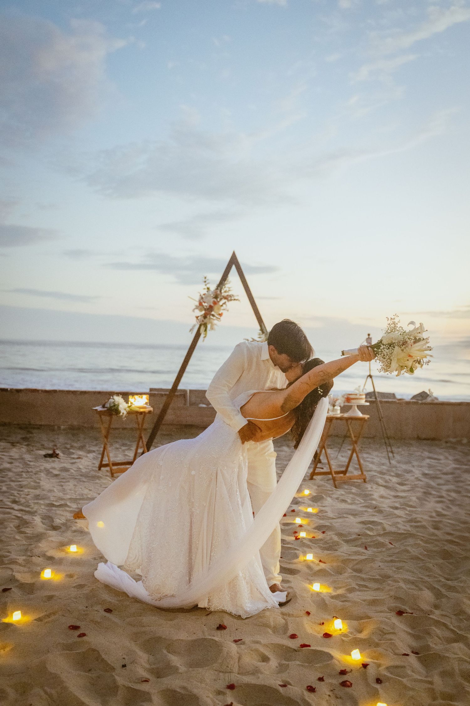 Bride in white dress playing guitar under triangle arch with glowing lights on beach at sunset.