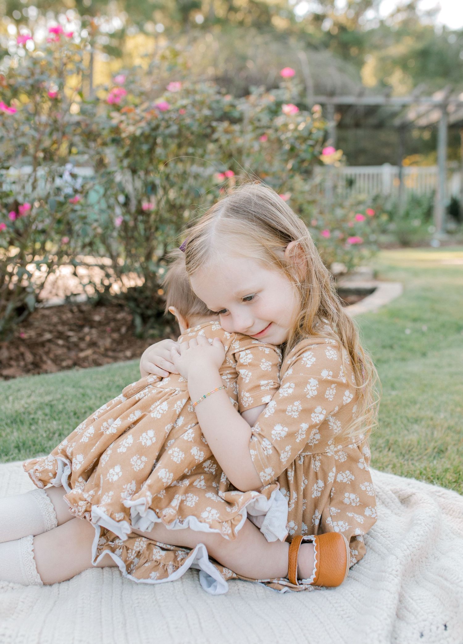 Young child in polka dot dress sitting on blanket in garden with blooming roses.
