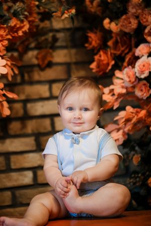 Baby in white outfit and blue suspenders sits against brick wall with orange flowers in background.