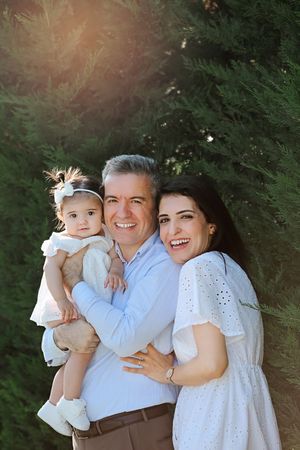 Family portrait in white outfits against green evergreen trees with sunlight peeking through.