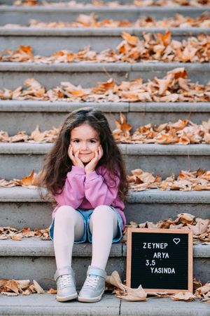 Child sitting on autumn-covered steps next to a black letter board sign.