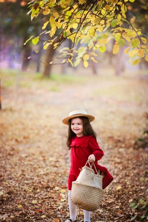 Child in red dress and straw hat carries woven basket through autumn forest with golden leaves.