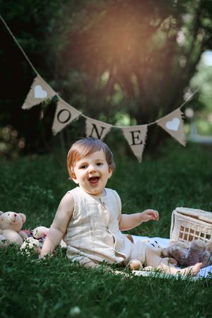 A joyful first birthday celebration outdoors with 'ONE' banner and teddy bears on a blanket in the grass.