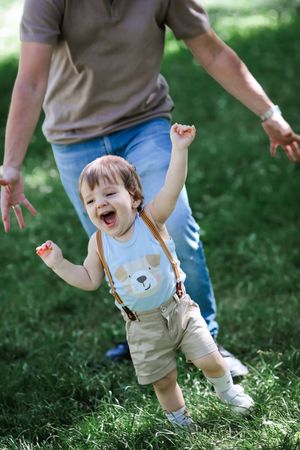 A happy baby in a bear-themed outfit runs excitedly through green grass outdoors.