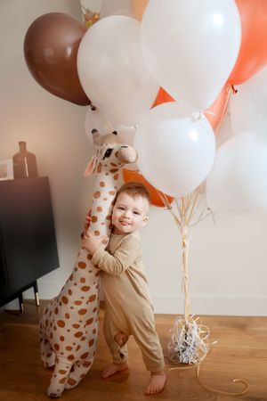 A toddler hugging a large giraffe plush toy surrounded by colorful balloons in a living room.