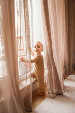 A curious toddler in beige outfit exploring sheer curtains by a window.