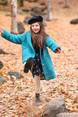 Child wearing bright blue coat and beret plays in fallen autumn leaves.