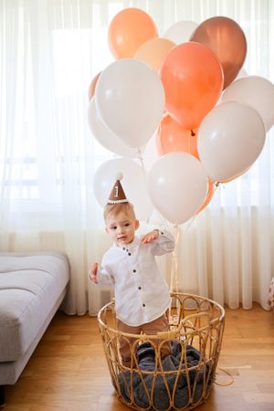 A toddler in party hat stands in wicker basket with orange and white balloons.