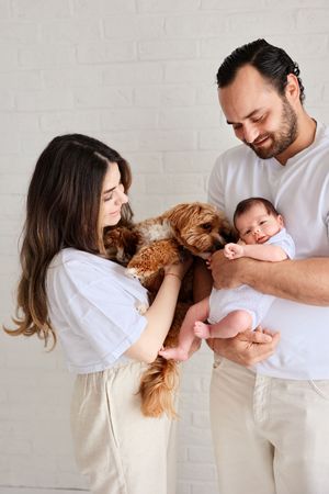 Family moment with goldendoodle dog and newborn baby against white brick wall.