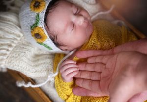 Peaceful newborn wearing yellow knit outfit and sunflower hat resting on soft white fabric.
