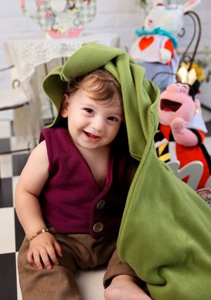 Playful toddler wearing burgundy vest and green cape smiling at camera with toys in background.