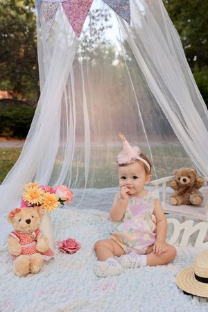 Baby sitting under tulle canopy with teddy bears and floral decorations.