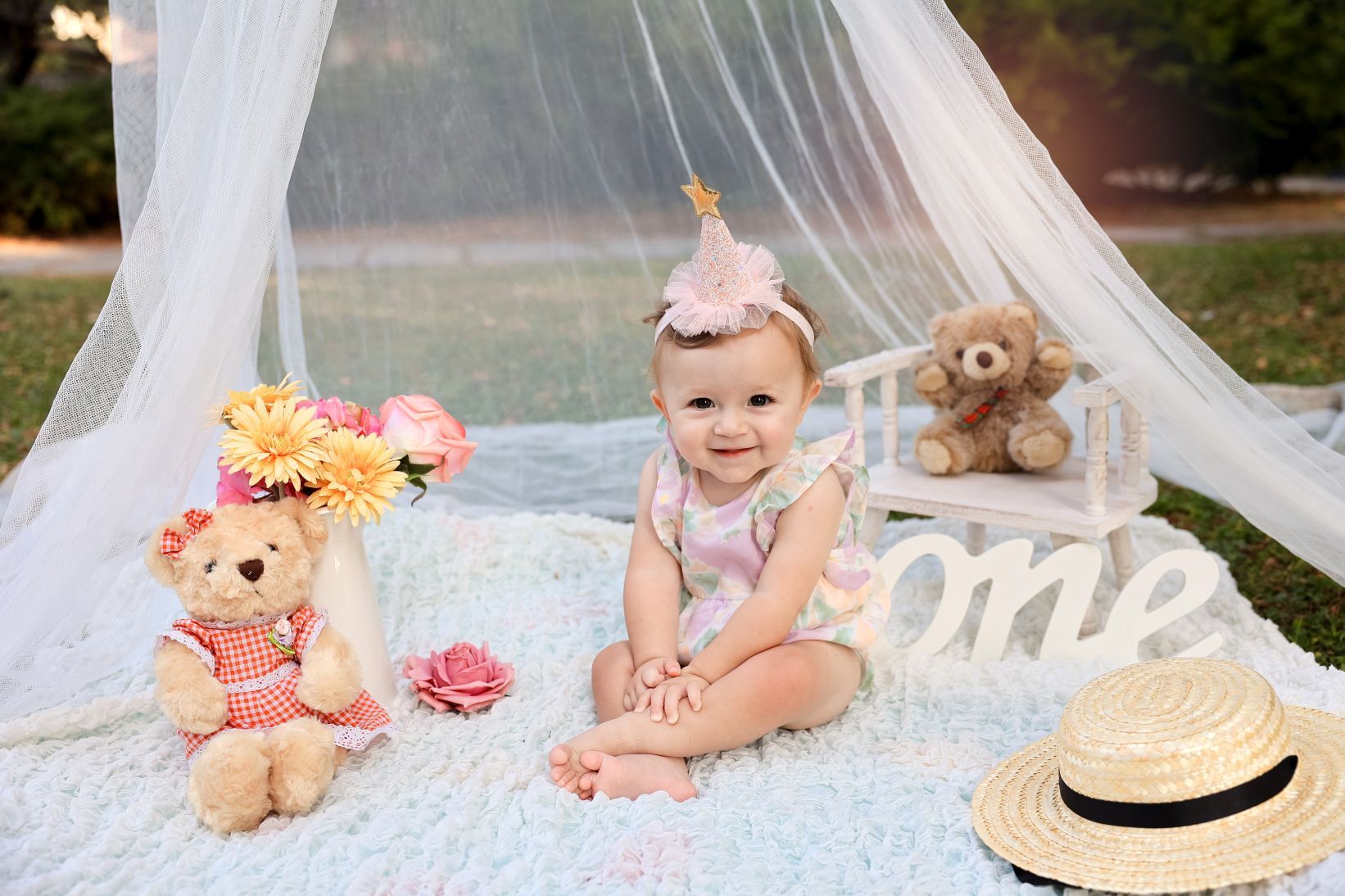 Baby sitting on blanket under sheer canopy with teddy bears and straw hat.