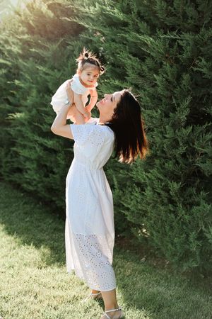 Mother in white dress lifting baby outdoors against cypress trees in summer sunshine.