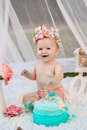 Baby wearing floral crown smiling with messy turquoise smash cake.