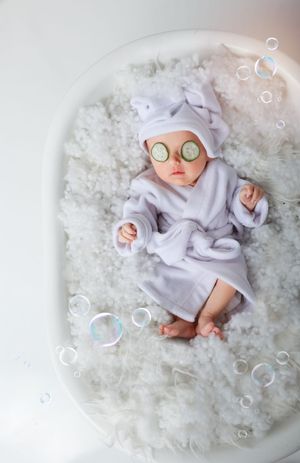 Creative newborn photo with baby in spa robe and cucumber slices surrounded by bubbles.