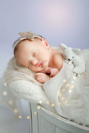 Sleeping newborn in white blanket with sparkly star headband and plush bunny toy amid twinkling fairy lights.