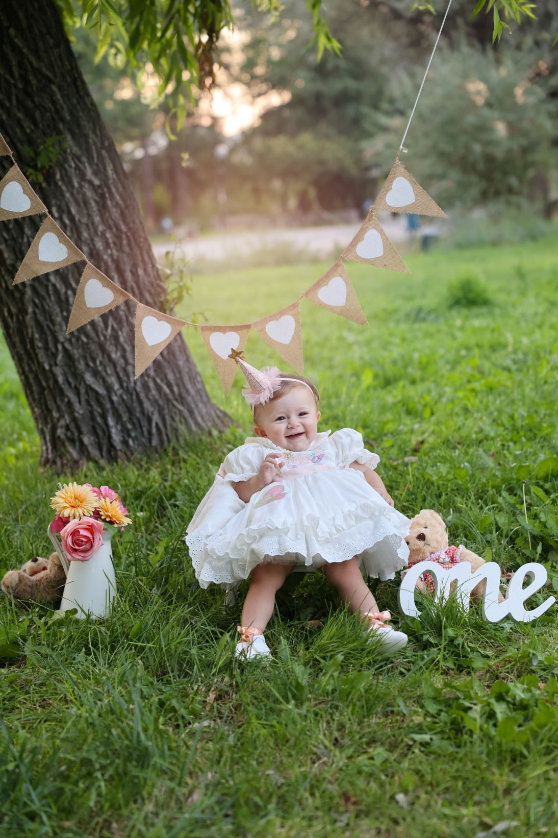 Baby in white dress sitting on grass under bunting decorations with floral arrangements.