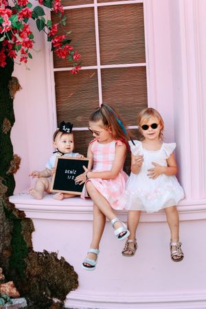 Three children in dresses sitting on pink window ledge with red flowers and bamboo blinds.