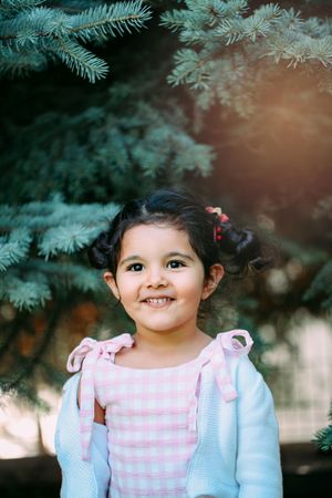 Young child in pink checkered dress and blue cardigan standing by evergreen branches.