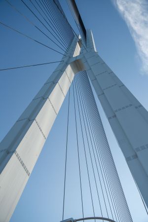Dramatic upward view of white suspension bridge cables and tower against blue sky.