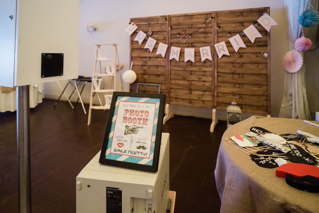 An indoor photo booth setup with a rustic wooden pallet backdrop, a "PHOTO BOOTH" sign on a stand, and a table full of props.