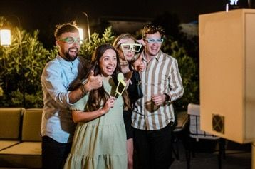 Four happy women posing in a photo booth with fun props and a rustic wooden background featuring a "LOVE" sign.