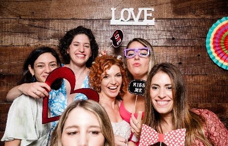 Six happy women posing in a photo booth with fun props and a rustic wooden background featuring a "LOVE" sign.
