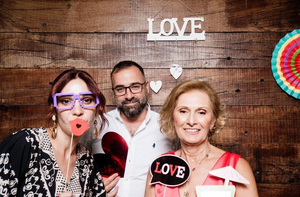 Three guests posing for a fun photo booth picture in front of a rustic wooden backdrop with "LOVE" signs and party props.
