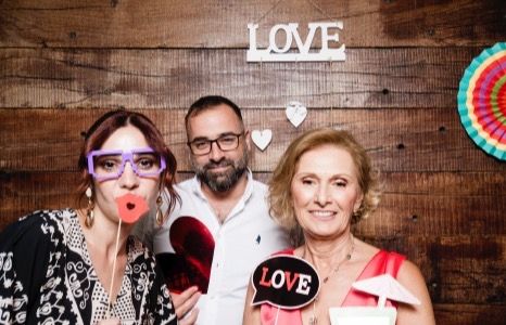 Three guests posing for a fun photo booth picture in front of a rustic wooden backdrop with "LOVE" signs and party props.