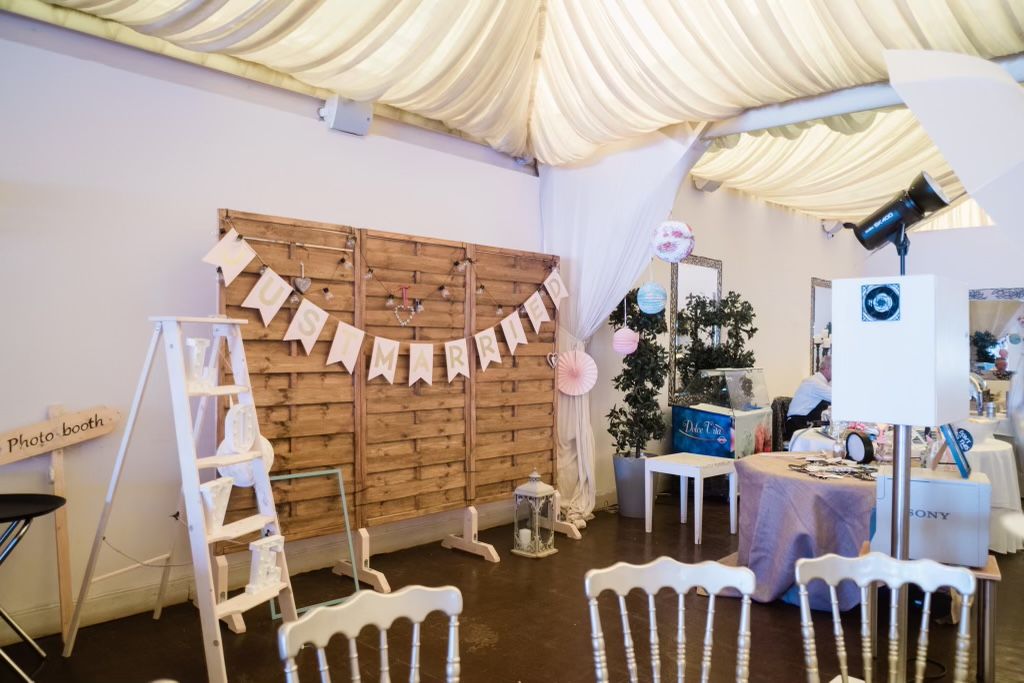 An indoor rustic event setup featuring a wooden pallet backdrop with a white bunting banner, vintage white chairs, and an open-air photo boo