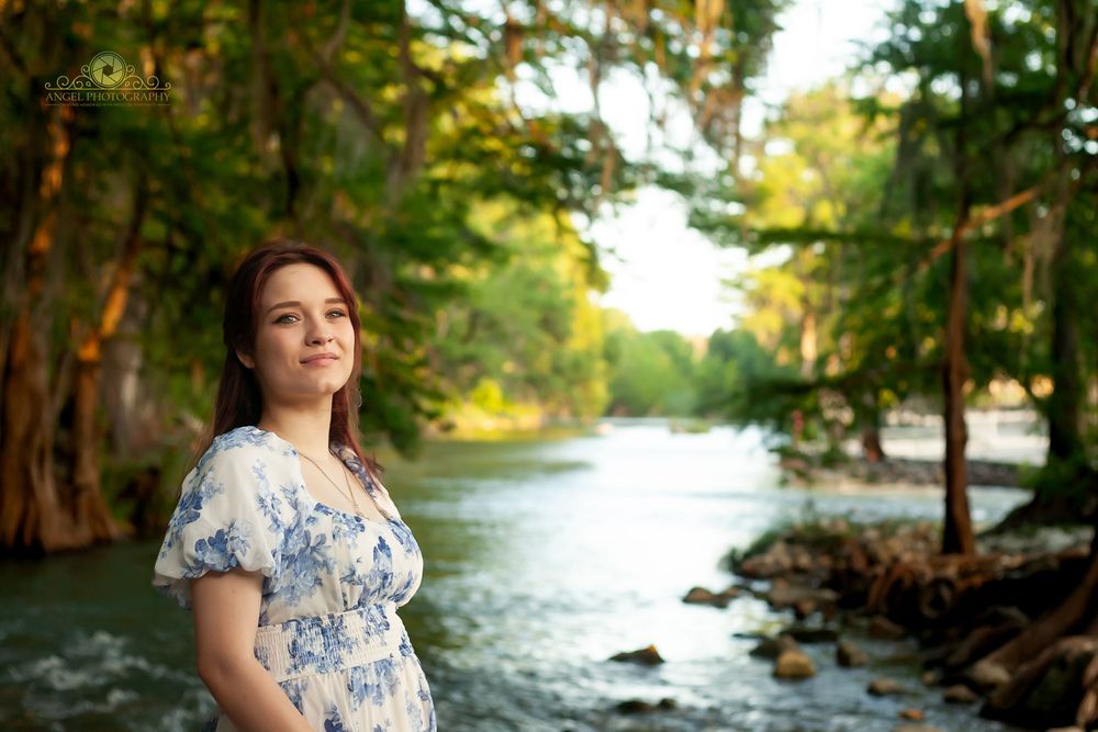Person in a floral dress stands peacefully by a flowing river surrounded by lush trees in warm sunlight.