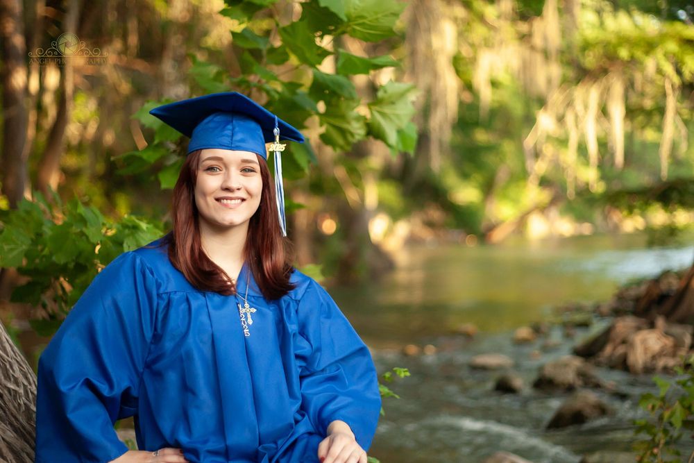 Graduate in bright blue cap and gown poses for photos outdoors in a natural wooded setting.