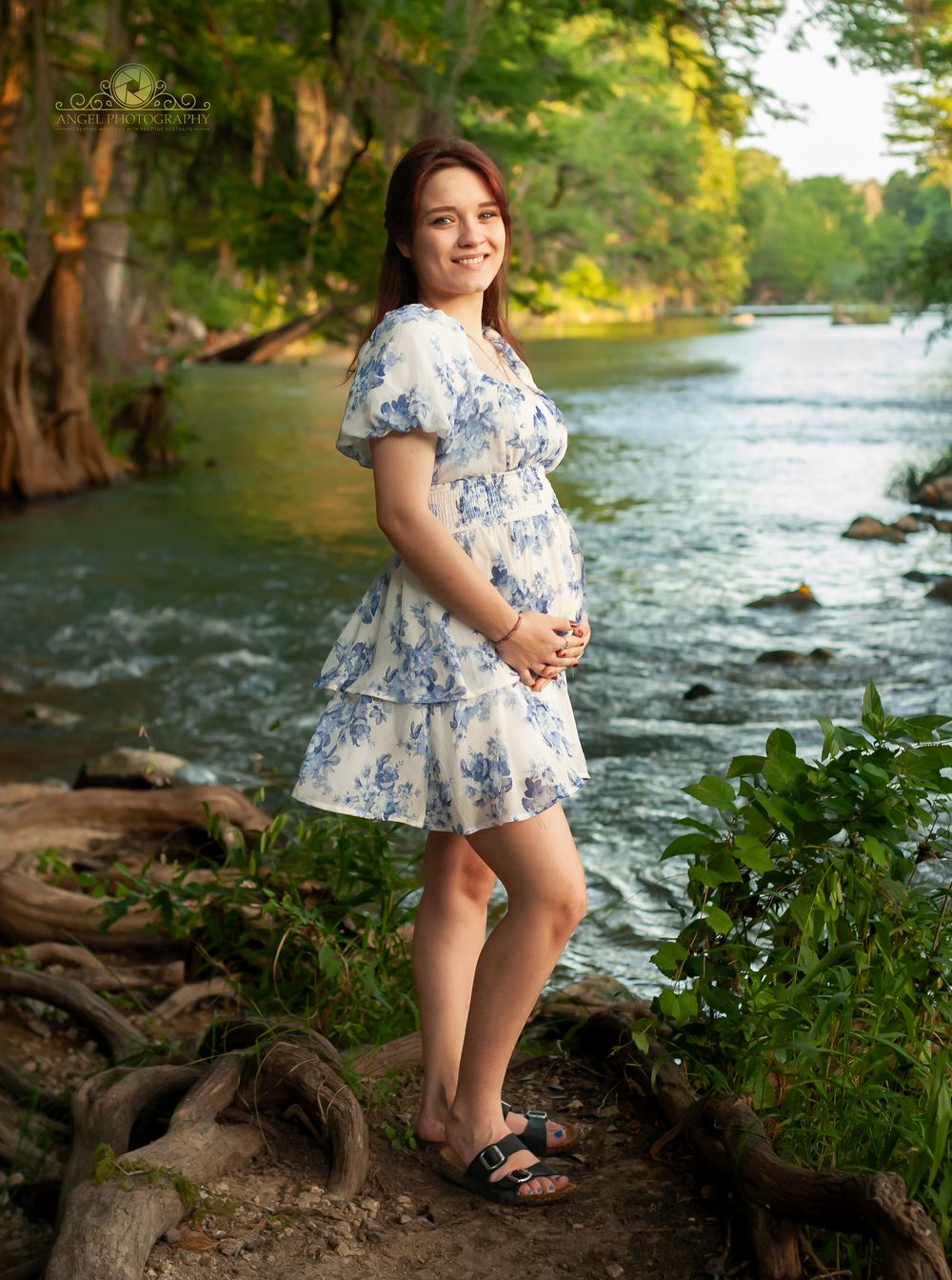 A child in a floral dress stands by a flowing river surrounded by trees and greenery during golden hour.