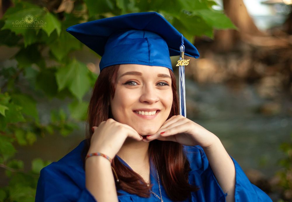 Graduate in blue cap and gown poses outdoors with hand resting on chin while smiling in natural setting.