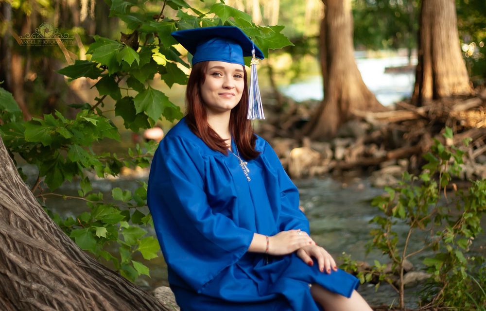 Graduate in blue cap and gown poses outdoors among trees during a sunny graduation photo session.