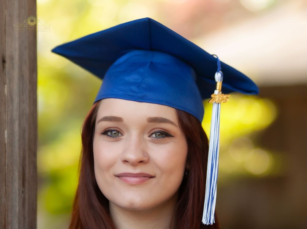 Graduate wearing blue cap and gown with white tassel smiles outdoors on sunny day.