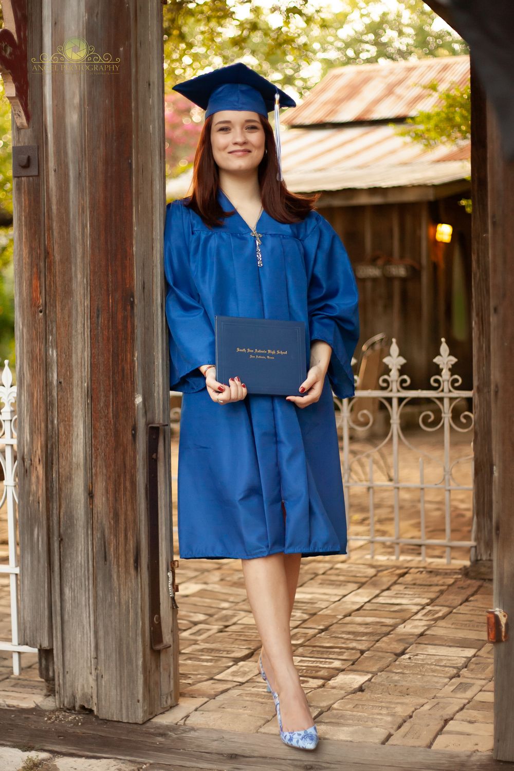 Graduate in blue cap and gown holds diploma while standing on rustic wooden porch with decorative iron gate.
