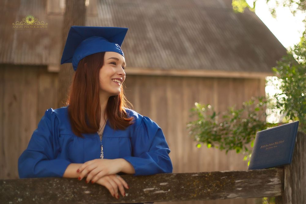 Graduate in blue cap and gown smiles while leaning on rustic wooden fence with barn in background.
