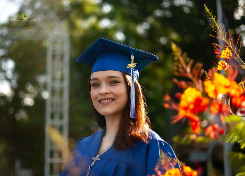 Graduate in blue cap and gown poses outdoors with vibrant orange flowers in the background.