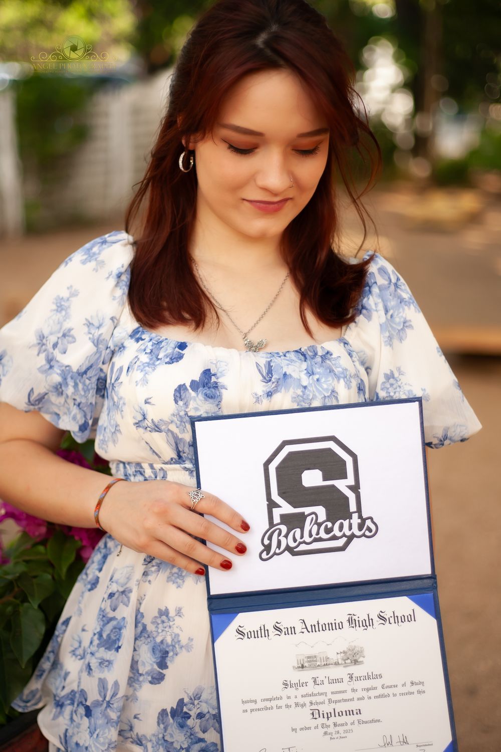 Graduate in blue and white floral dress holds diploma and school logo from South San Antonio High School.