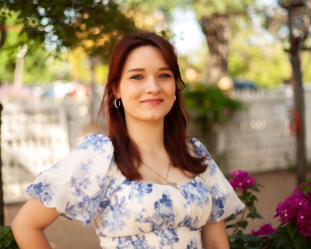 A woman in a floral dress poses outdoors near blooming flowers in warm natural lighting.