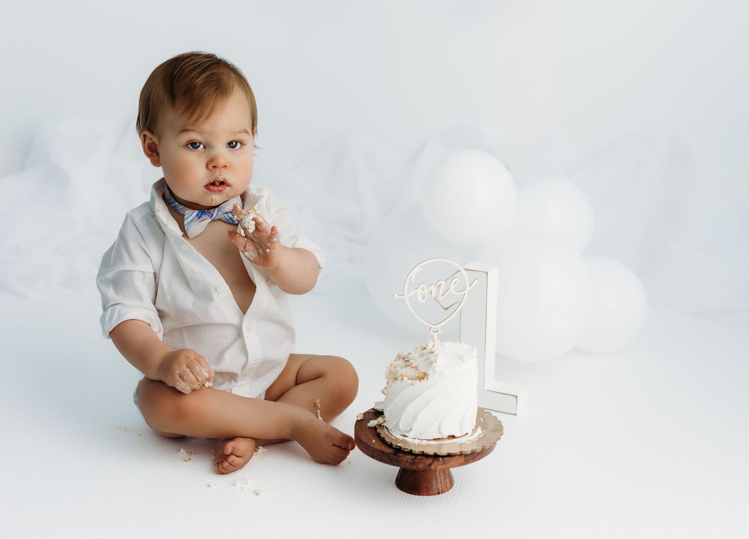 Baby in white shirt sits next to a rustic cake stand with white cake and number one candle for first birthday photo.
Rashinphotography-Nova